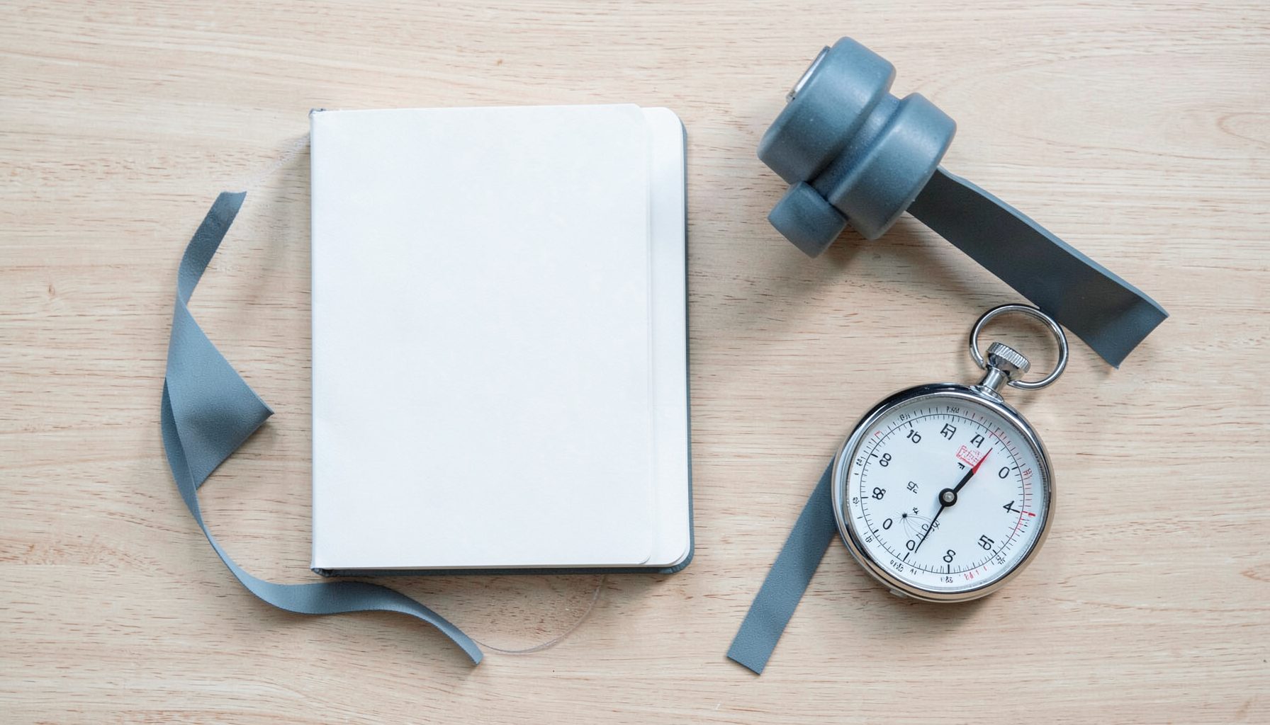 A clean overhead view of a notebook, dumbbell, resistance band, and stopwatch arranged on a light wood surface, with soft natural light and muted steel-blue accents