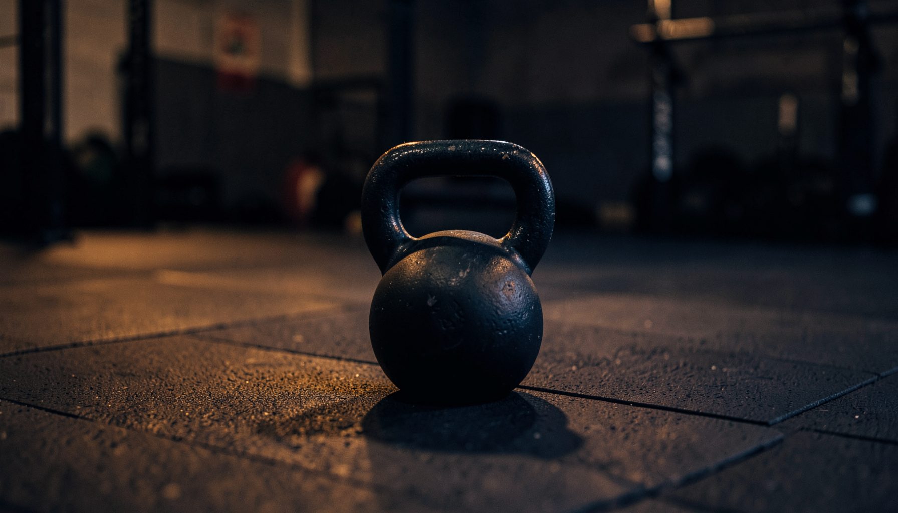 A dimly lit gym floor with a single kettlebell resting on rubber matting, warm amber and deep navy tones, calm and focused atmosphere.