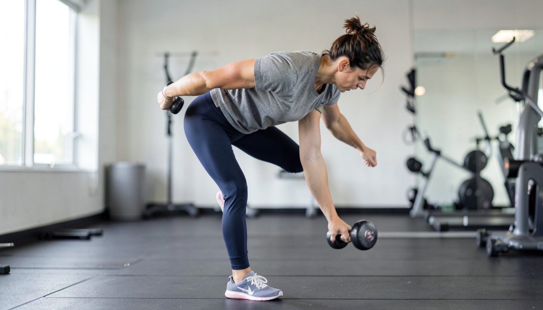 A person balancing on one leg while hinging forward with a dumbbell in the opposite hand, soft natural light, muted gray and navy workout attire, clean gym backdrop.