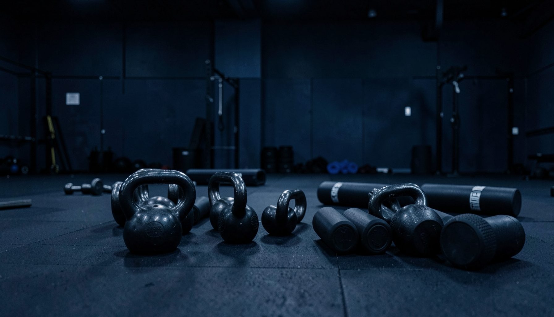 A dimly lit gym floor with kettlebells and foam rollers arranged neatly, deep navy and steel blue tones dominating the scene.