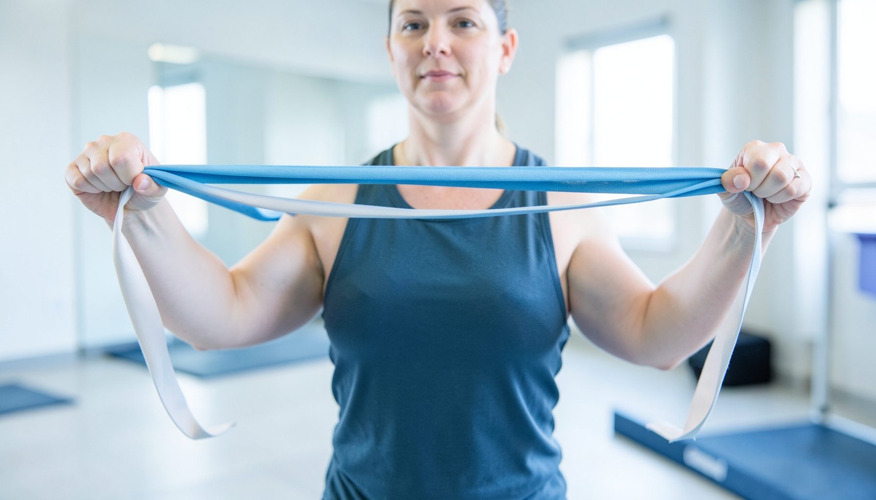 Resistance band stretched between two hands at shoulder height, soft blue and gray tones, bright clean training space.